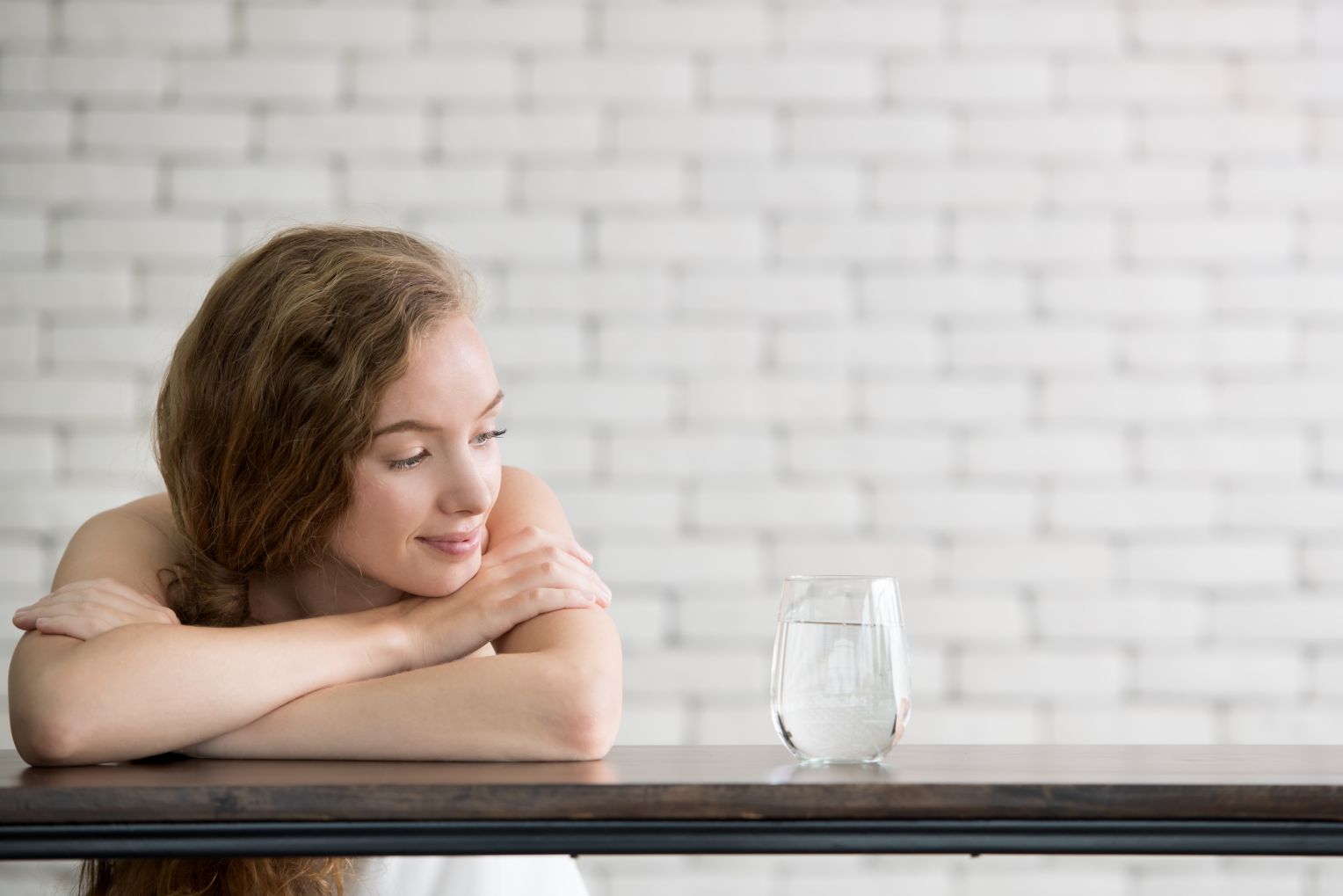 Eine junge Frau mit langen, lockigen Haaren sitzt entspannt an einem Tisch und blickt lächelnd auf ein Glas Wasser. Im Hintergrund ist eine weiße Ziegelwand zu sehen.