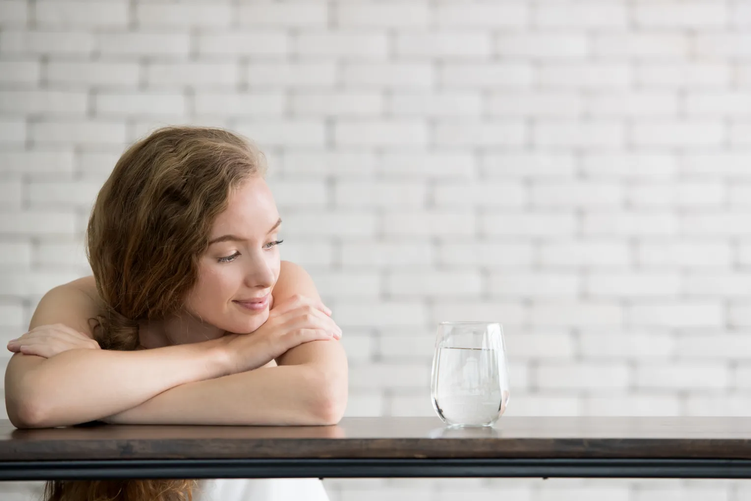 Eine junge Frau mit langen, lockigen Haaren sitzt entspannt an einem Tisch und blickt lächelnd auf ein Glas Wasser. Im Hintergrund ist eine weiße Ziegelwand zu sehen.