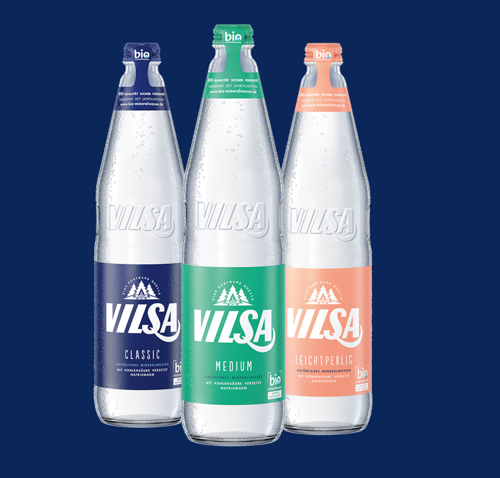 Drei Glasflaschen der Marke VILSA mit Mineralwasser in den Sorten Classic, Medium und Leichtperlig, jeweils mit unterschiedlich farbigen Etiketten (blau, grün, orange) vor dunkelblauem Hintergrund.