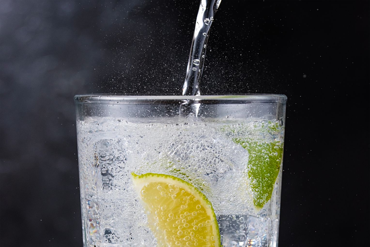 Ein Glas mit sprudelndem Mineralwasser, das mit Limettenscheiben und Eiswürfeln gefüllt ist. Frisches Wasser wird gerade ins Glas gegossen.