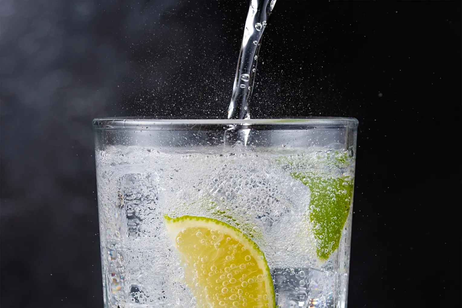 Ein Glas mit sprudelndem Mineralwasser, das mit Limettenscheiben und Eiswürfeln gefüllt ist. Frisches Wasser wird gerade ins Glas gegossen.
