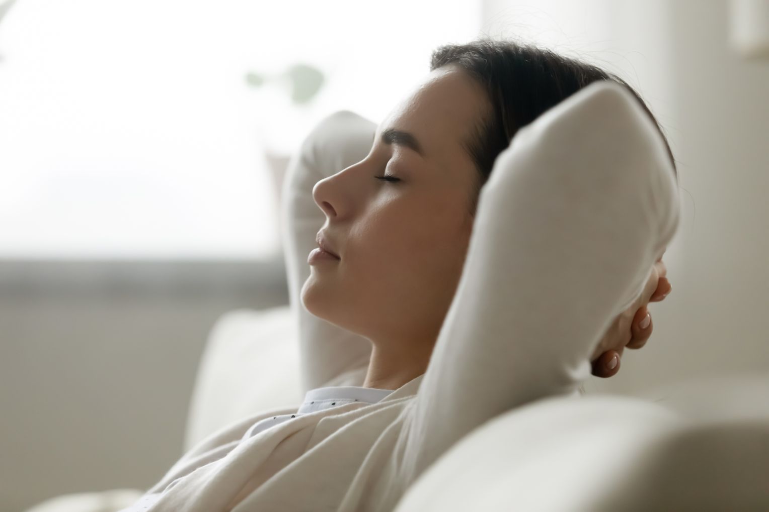 Eine junge Frau lehnt entspannt auf einem Sofa mit geschlossenen Augen und verschränkten Armen hinter dem Kopf. Sie wirkt ruhig und ausgeglichen in einem hellen, gemütlichen Raum.