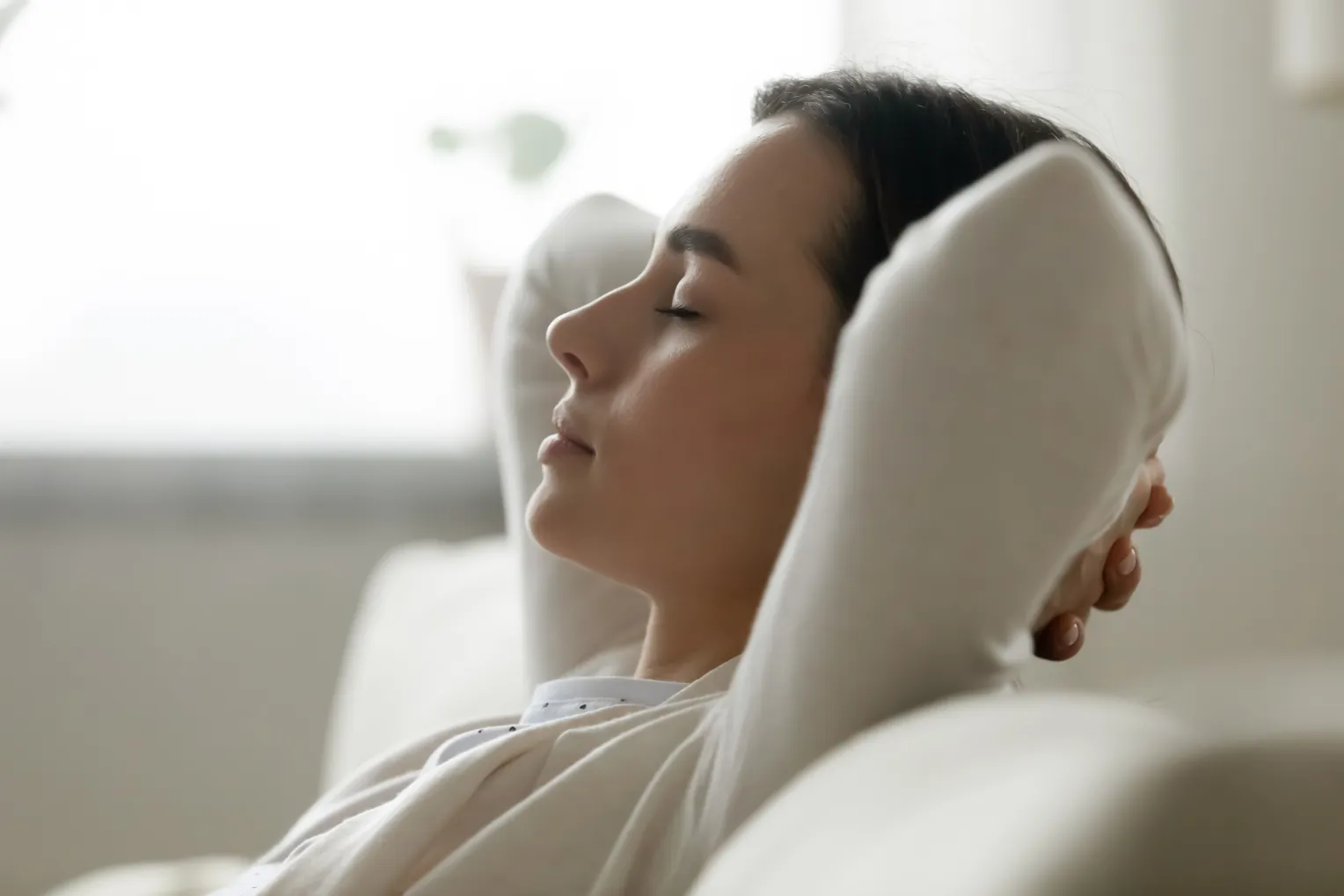 Eine junge Frau lehnt entspannt auf einem Sofa mit geschlossenen Augen und verschränkten Armen hinter dem Kopf. Sie wirkt ruhig und ausgeglichen in einem hellen, gemütlichen Raum.