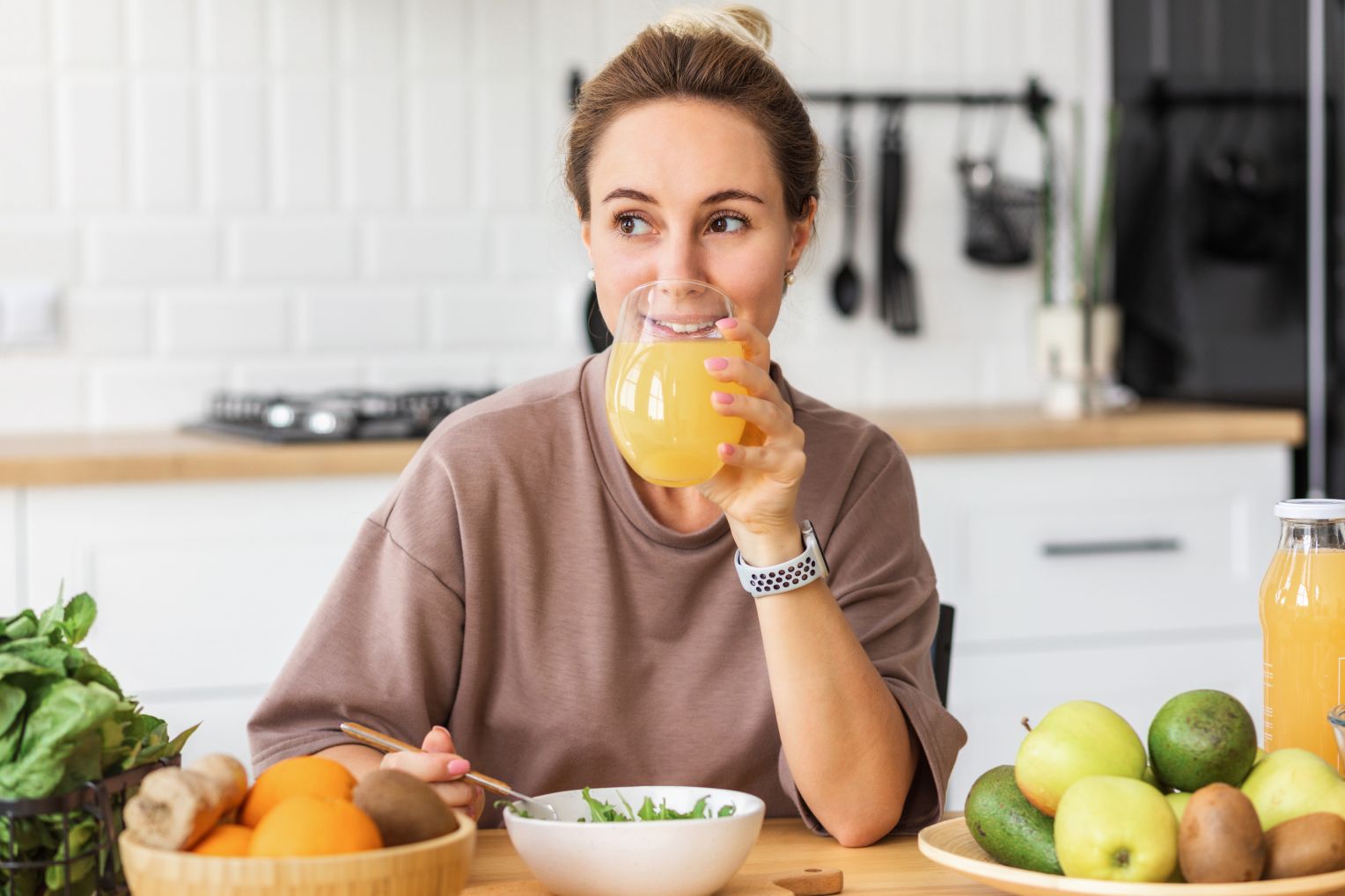 Eine lächelnde Frau sitzt an einem Tisch in einer modernen Küche, trinkt ein Glas Orangensaft und isst einen Salat. Auf dem Tisch stehen frisches Obst, Gemüse und eine Flasche Saft.