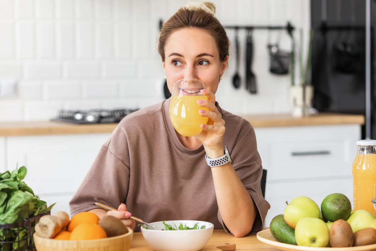 Eine lächelnde Frau sitzt an einem Tisch in einer modernen Küche, trinkt ein Glas Orangensaft und isst einen Salat. Auf dem Tisch stehen frisches Obst, Gemüse und eine Flasche Saft.