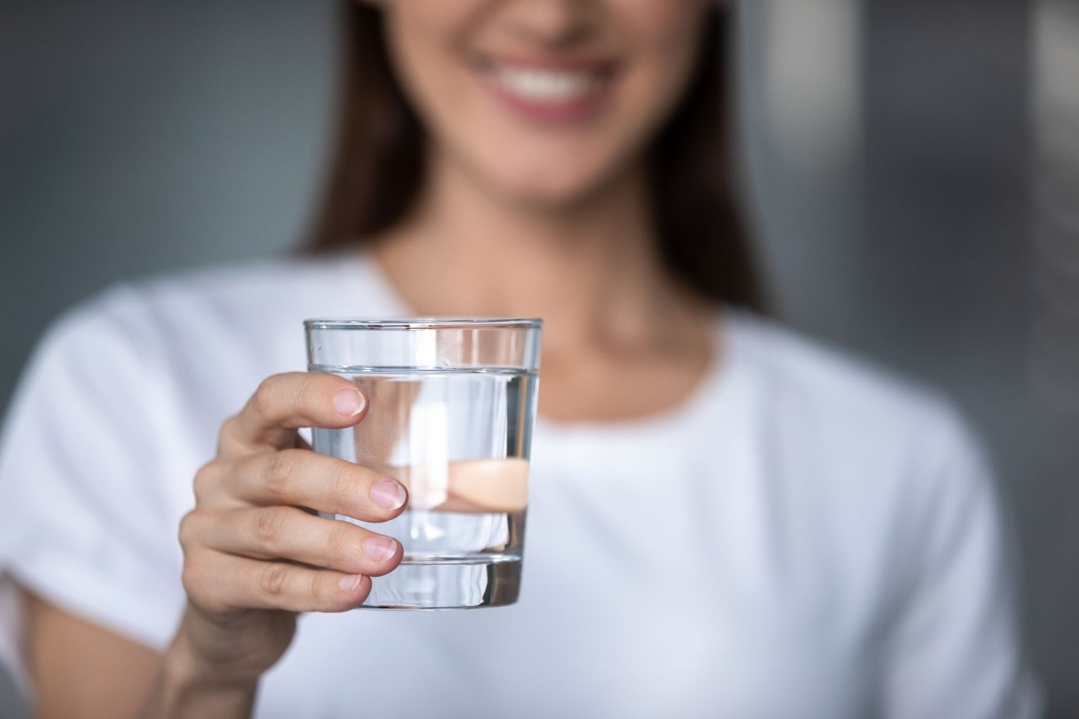 Eine Frau in einem weißen T-Shirt hält ein Glas klares Wasser in die Kamera. Ihr Gesicht ist nur teilweise sichtbar, sie lächelt.