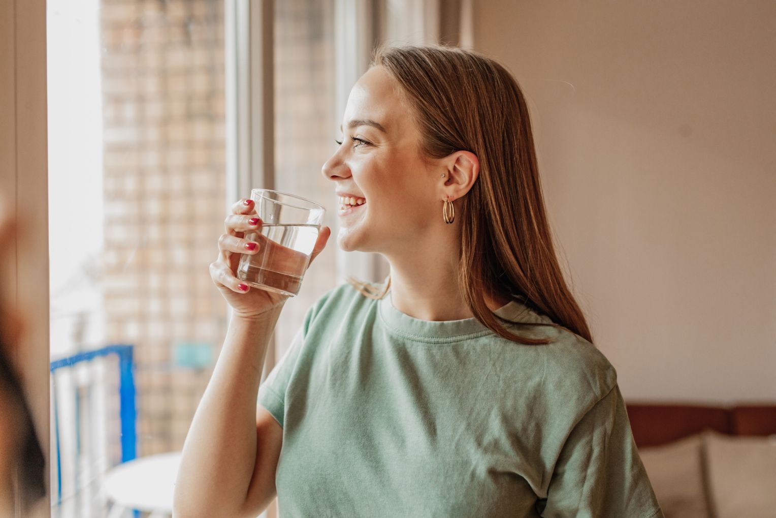 Lächelnde junge Frau steht am Fenster und hält ein Glas Wasser in der Hand.
