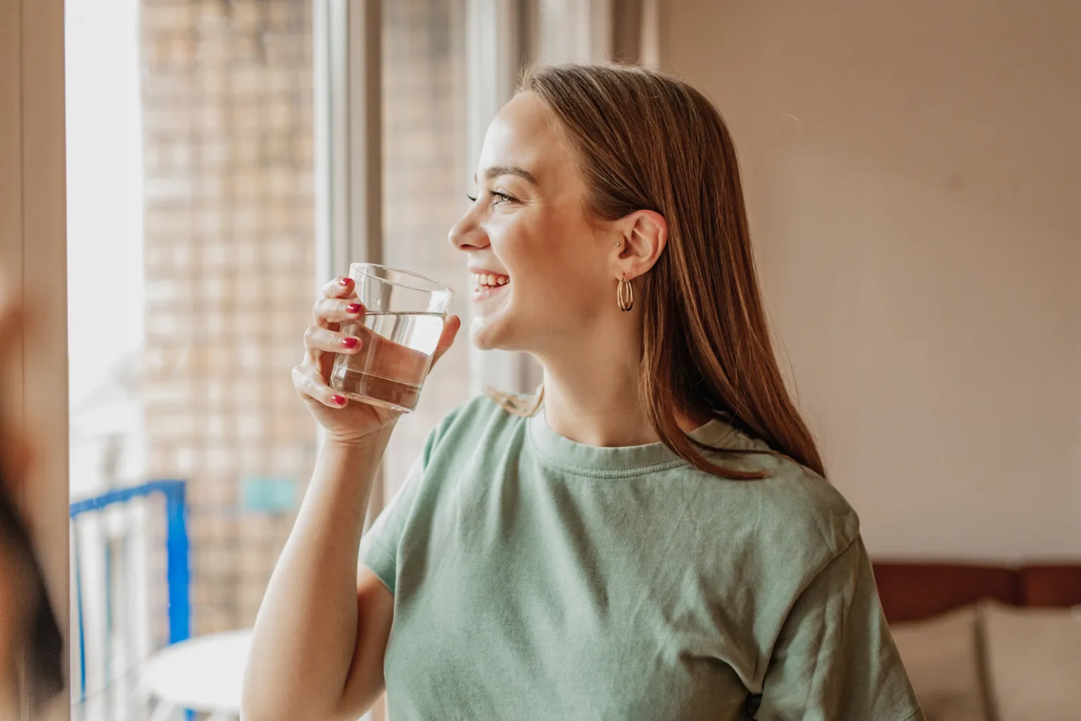 Lächelnde junge Frau steht am Fenster und hält ein Glas Wasser in der Hand.