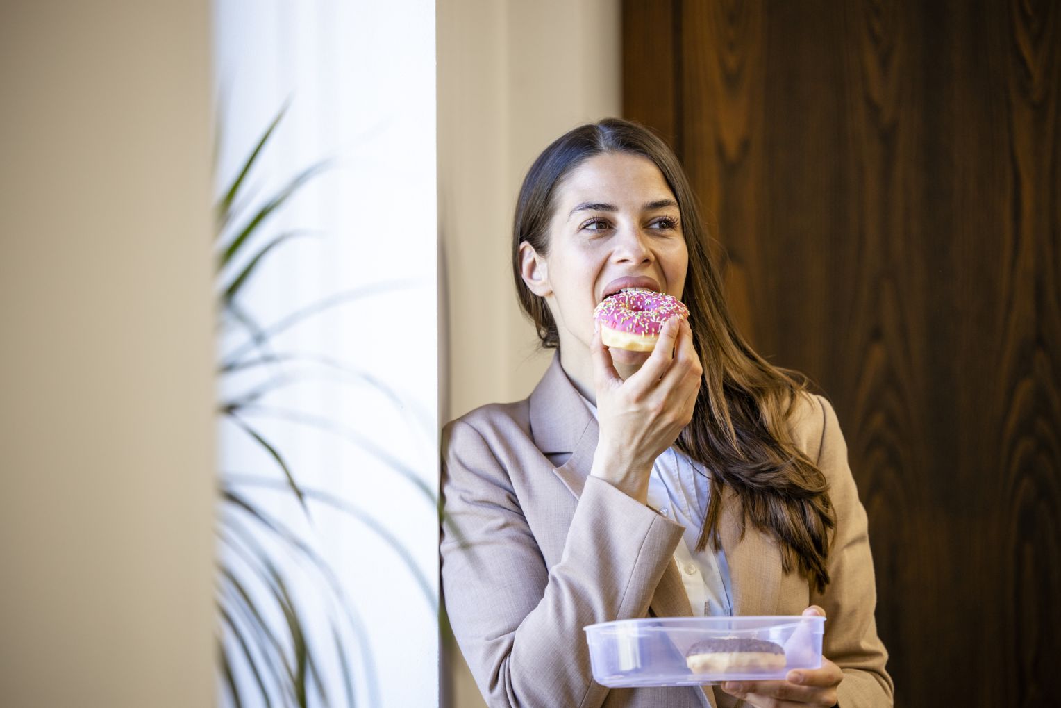 Frau in heller Jacke beißt in einen rosa Donut mit bunten Streuseln und hält eine Plastikbox mit weiteren Donuts.