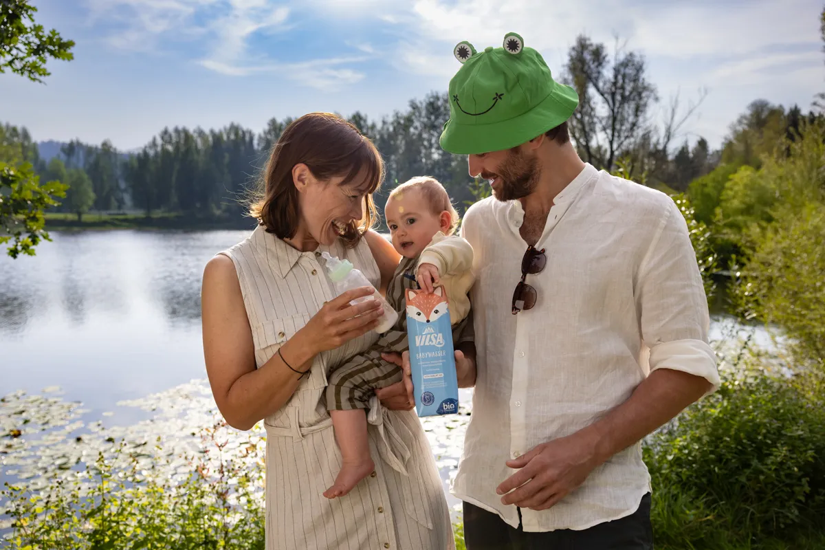 Eine Frau und ein Mann stehen mit einem Kleinkind am See. Der Mann hält eine geöffnete VILSA Tetra Babywasser Packung in der Hand.
