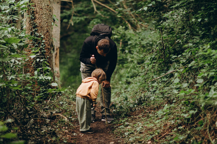 Eine erwachsene Person mit Rucksack beugt sich auf einem schmalen Waldweg zu einem kleinen Kind hinunter; beide sind von dichtem grünen Wald umgeben und scheinen gemeinsam den Boden zu erkunden.