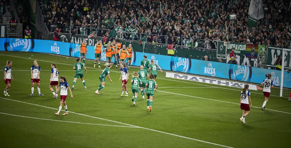 Spielszene im Frauenfußball vor einer vollen Fankurve, mit sichtbarer VILSA-Werbebande am Spielfeldrand.
