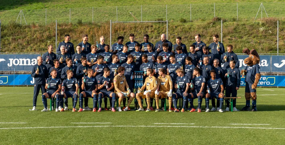 Fußballmannschaft und Betreuerteam posieren auf dem Trainingsplatz mit VILSA-Getränken und einem Maskottchen für ein Gruppenfoto.
