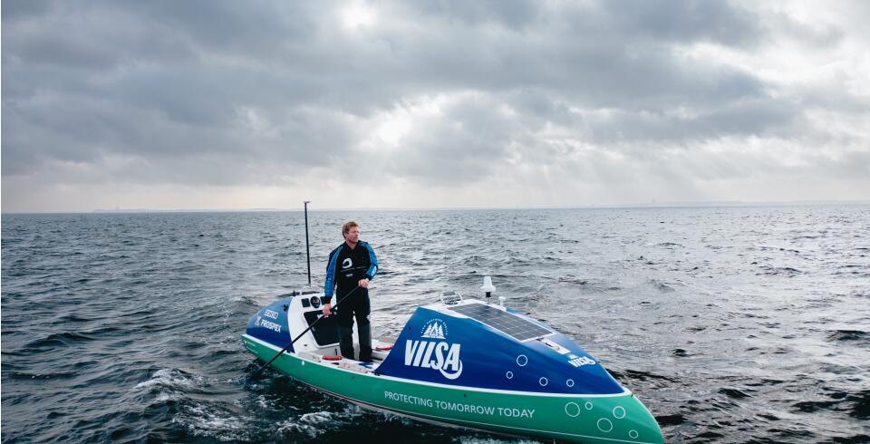 Ruderer steht in einem VILSA-gebrandeten Boot auf offenem Wasser unter bewölktem Himmel.
