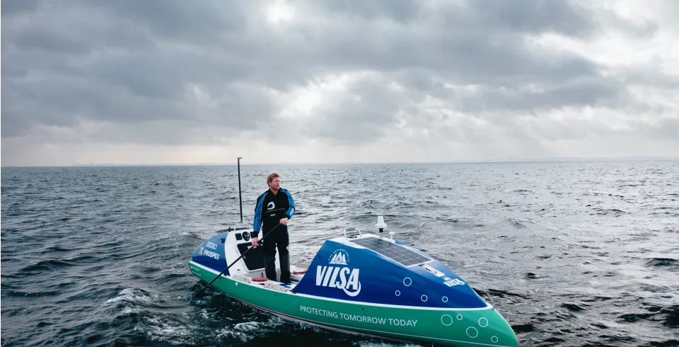 Ruderer steht in einem VILSA-gebrandeten Boot auf offenem Wasser unter bewölktem Himmel.