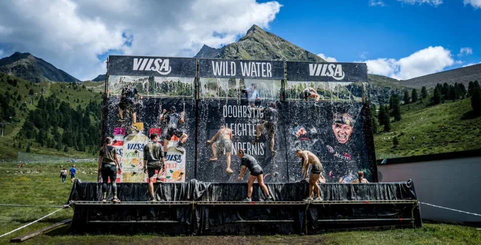 Teilnehmende überwinden beim Hindernislauf ein VILSA-gebrandetes „Wild Water“-Wasserhindernis in den Bergen.