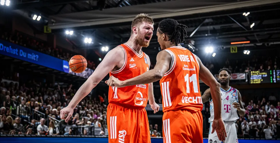 Basketballspieler in orangefarbenen Trikots jubeln während eines Spiels in einer Halle; auf der Hose und an der Bande ist VILSA-Werbung zu sehen.