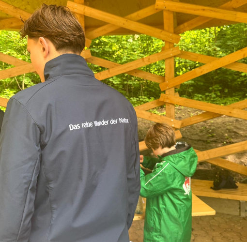 Person mit Jacke und Aufschrift „Das reine Wunder der Natur“ steht mit einem Kind in einer Holzhütte im Grünen.