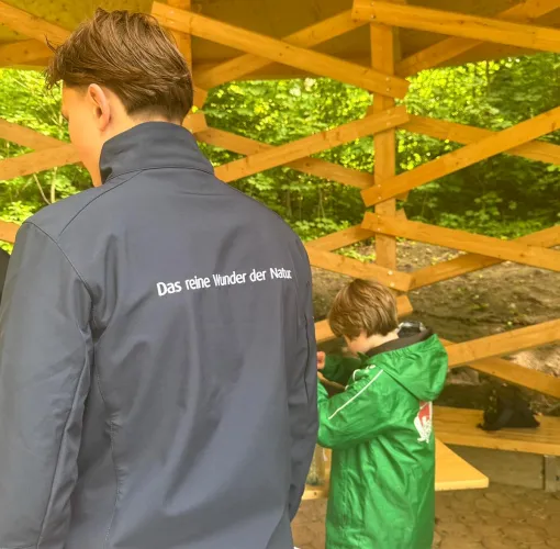 Person mit Jacke und Aufschrift „Das reine Wunder der Natur“ steht mit einem Kind in einer Holzhütte im Grünen.