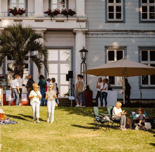 Menschen sitzen und spazieren auf einer Wiese vor einem historischen Gebäude bei spätnachmittäglichem Licht.