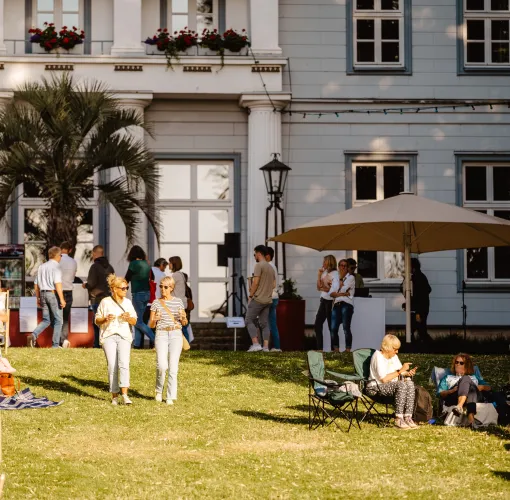 Menschen sitzen und spazieren auf einer Wiese vor einem historischen Gebäude bei spätnachmittäglichem Licht.