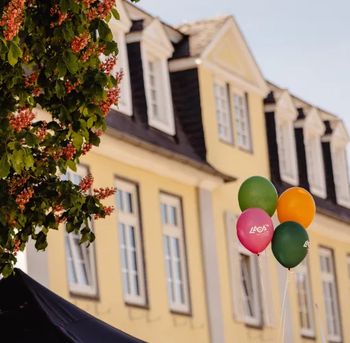 Bunte Luftballons mit LaGa-Logo schweben vor dem gelben Haus Kassel und blühenden Bäumen.