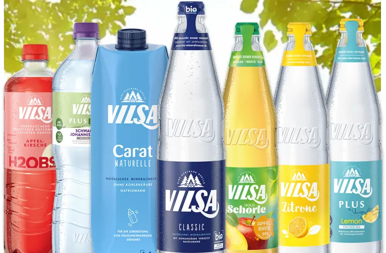 Mehrere VILSA-Getränkeflaschen und -packungen in einer Reihe auf einem Holztisch. Im Hintergrund sonniger, grüner Blätterhimmel. Sorten: VILSA H2Obst, PLUS Bio, Carat Naturelle, Classic Mineralwasser, Apfel-Schorle, Zitrone, und Lemon Calcium. Die Verpackungen sind bunt gestaltet in Rot, Lila, Blau, Grün und Gelb.