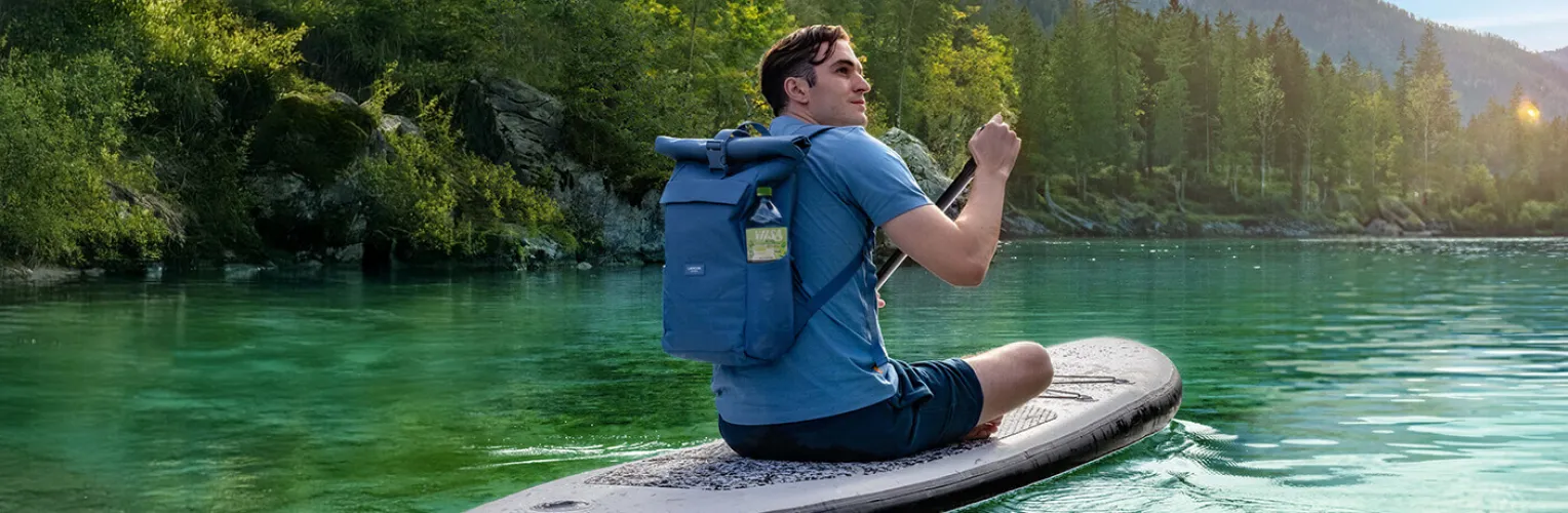 Mann sitzt auf einem Stand-up-Paddleboard auf einem klaren Bergsee; in der Seitentasche seines Rucksacks steckt eine Flasche VILSA PLUS Bio Limette-Minze 1,0 l.