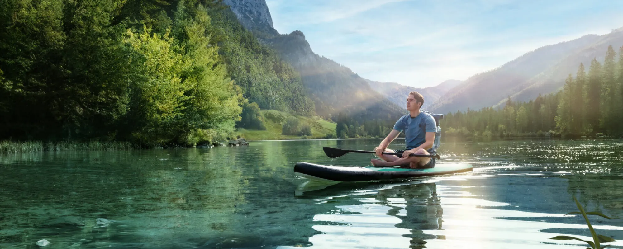 Mann sitzt auf einem Stand-up-Paddleboard auf einem klaren Bergsee, umgeben von bewaldeten Bergen und blickt in die Natur.