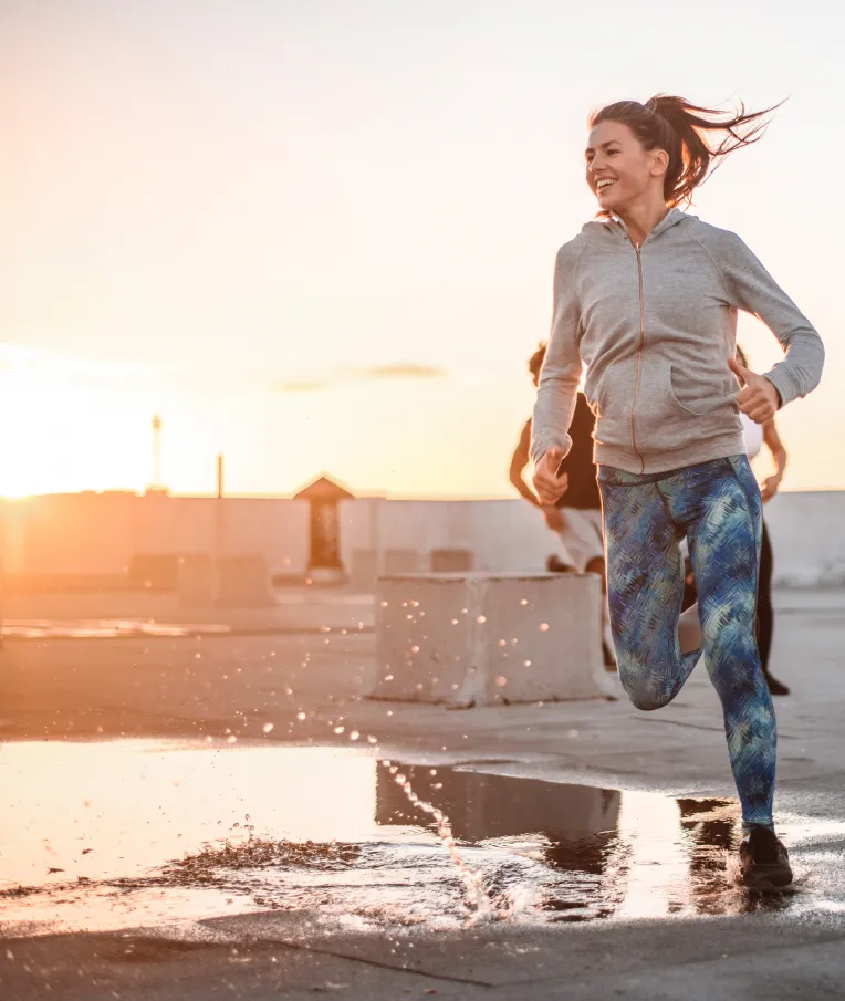 junge Frau mit grauer Jacke und blauer Sportleggings joggt im Sonnenschein durch eine Pfütze