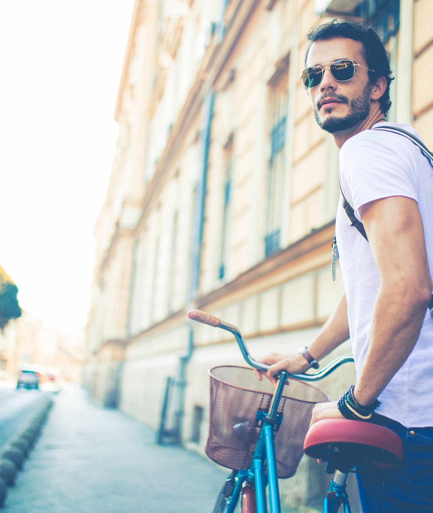 junger Mann mit dunklen Haaren, Sonnenbrille und helllila T-Shirt schiebt ein Fahrrad und guckt in die Ferne