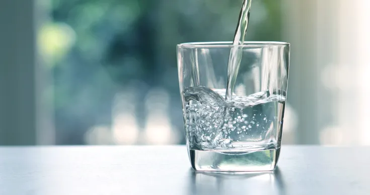 Ein klares Wasserglas steht auf einer hellen Oberfläche, während Wasser von oben hineingegossen wird. Im Glas sind Blasen und die Bewegung des Wassers sichtbar. Der Hintergrund ist weich, unscharf und in Blau- und Grüntönen.