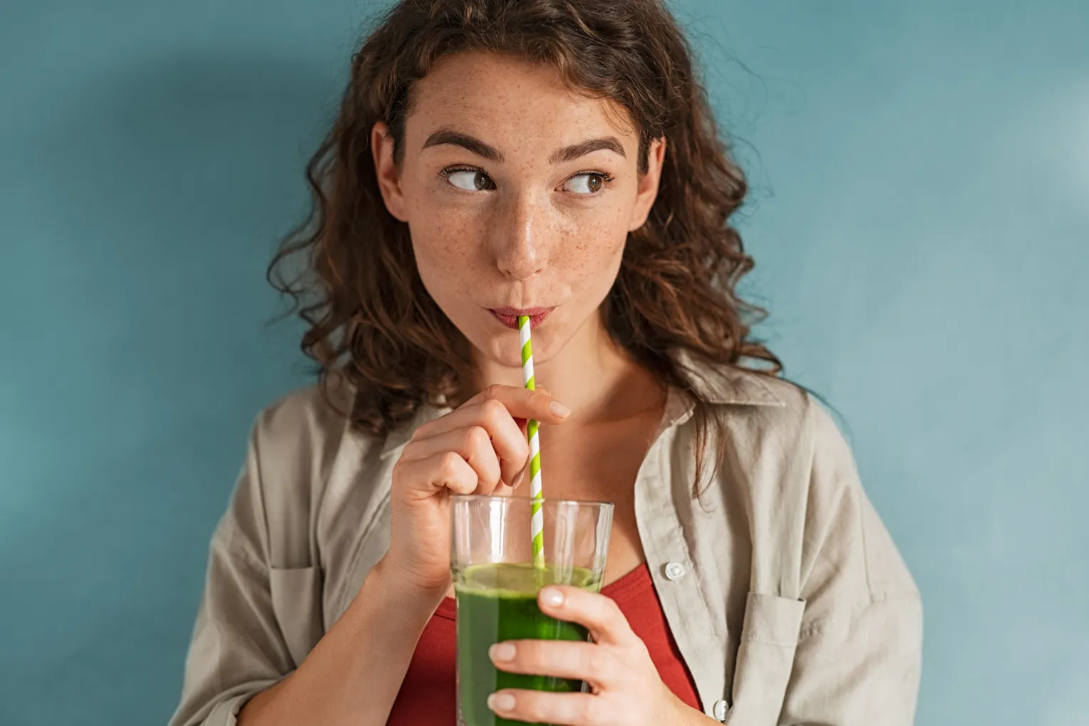 Eine Frau mit welligem, braunem Haar trinkt mit einem grün gestreiften Strohhalm aus einem Glas einen grünen Smoothie.