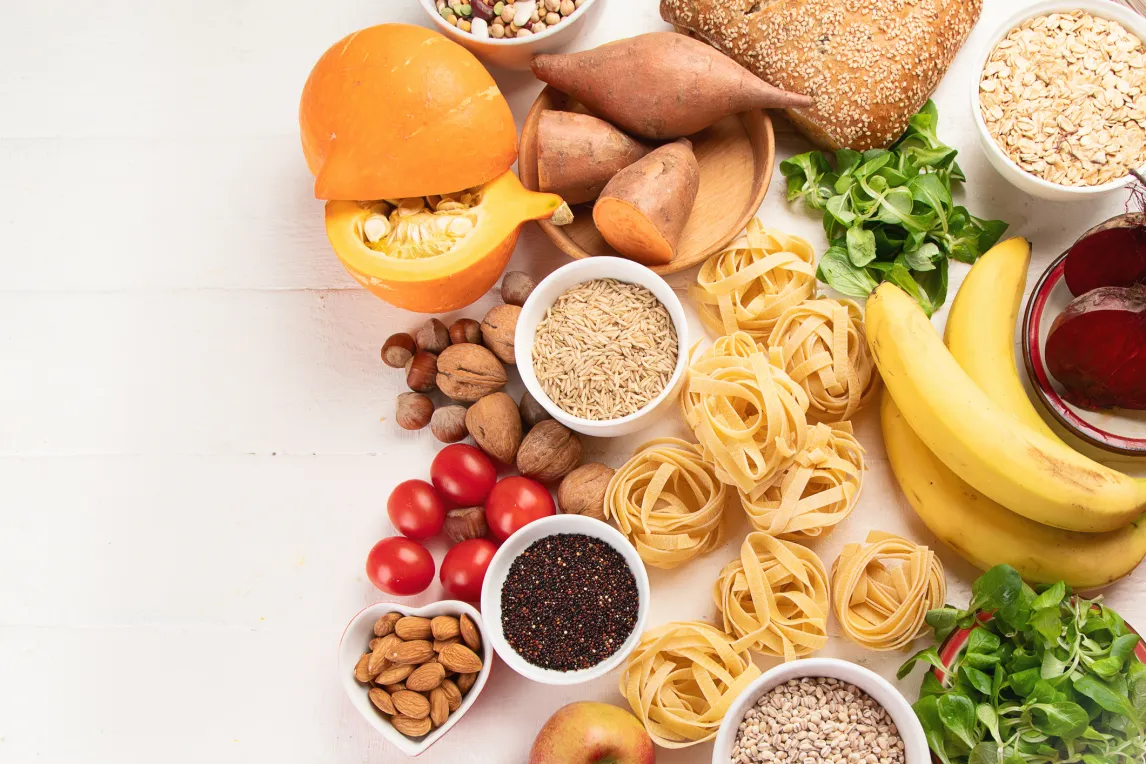 Auswahl aus Kürbis, Bananen, Tomaten, Nüssen, Getreide, Pasta und Blattgemüse auf hellem Untergrund.