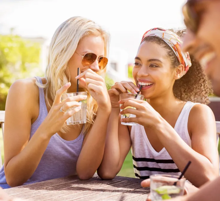 Zwei Frauen sitzen im Freien und trinken lächelnd ihre Kaltgetränken aus Gläsern.