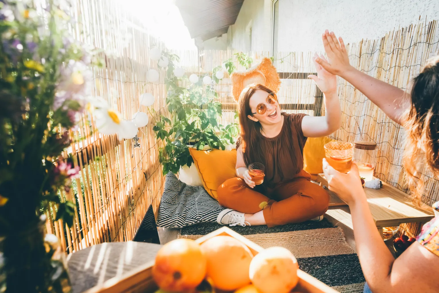 Zwei Personen stoßen auf einem sonnigen Balkon mit Getränken an und geben sich Highfive.