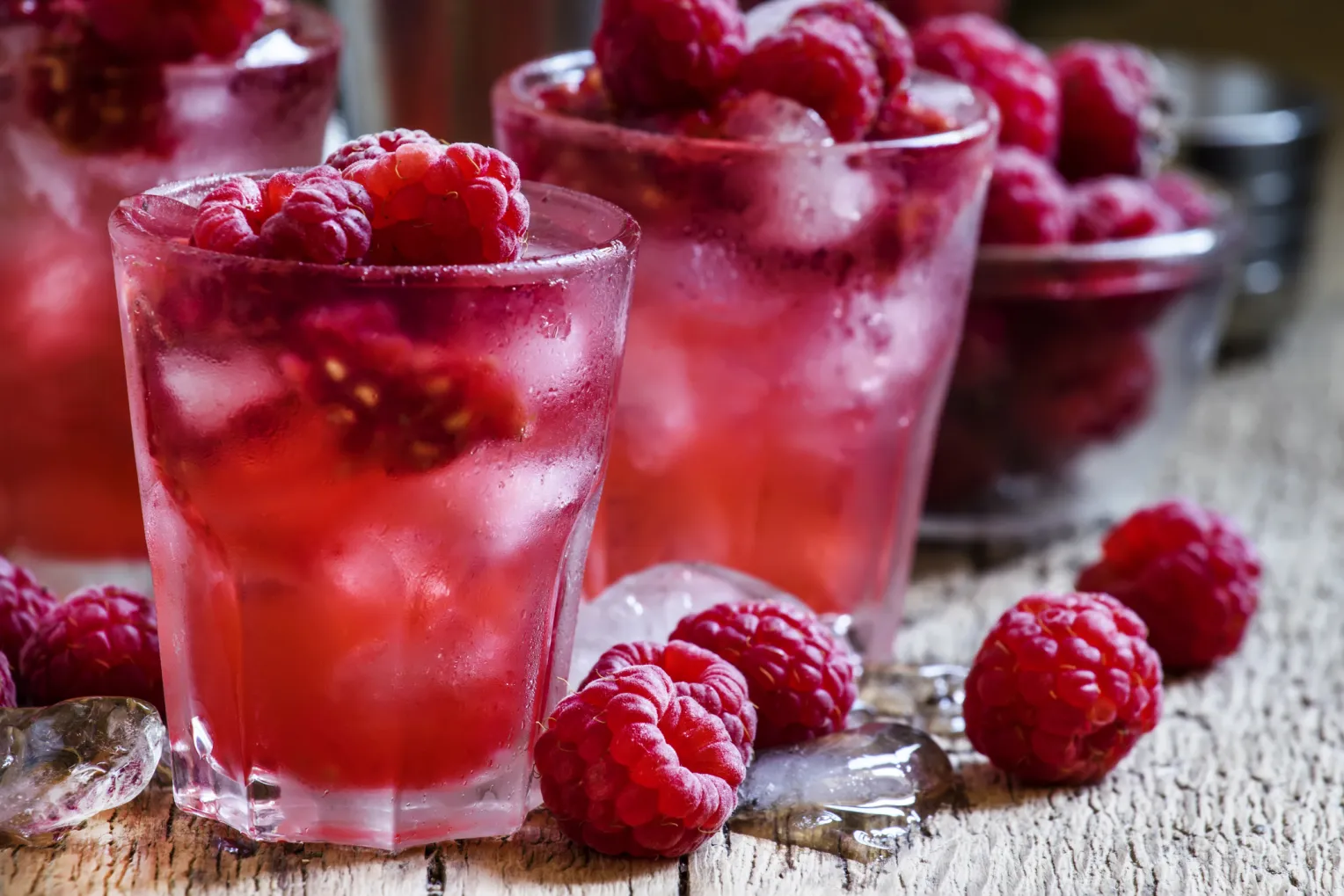 Nahaufnahme von drei Gläsern mit pinkem Kaltgetränk, Eiswürfeln und Himbeeren stehen auf einem Holztisch. Auf dem Tisch liegen vereinzelt auch Himbeeren und Eiswürfel neben den Gläsern.