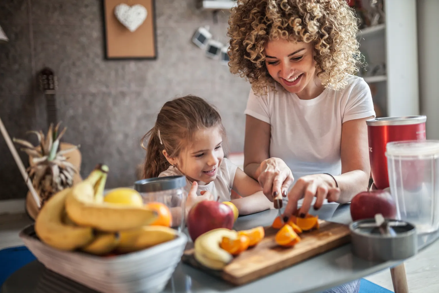 Eine Mutter und ihre Tochter schneiden gemeinsam Obst in einer Küche und bereiten einen Smoothie vor. Auf der Küchenablage sieht man verschiedene Arten von Obst: Bananen, Äpfel, Orangen.