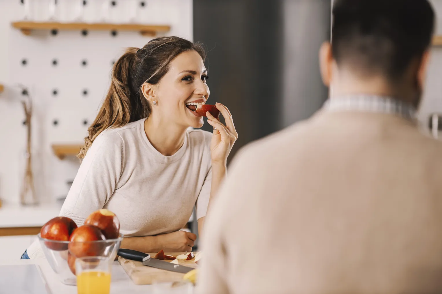Frau sitzt in einer Küche und isst ein Stück Apfel. Auf dem Tisch liegen weitere Äpfel und ein Glas Saft.