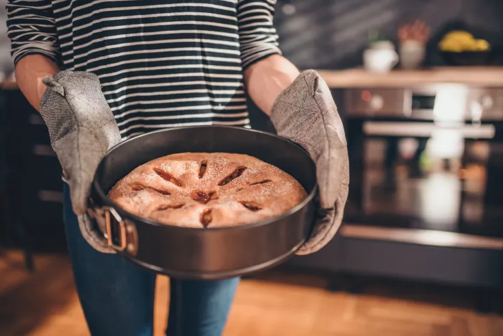 Person hält mit Ofenhandschuhen eine Backform mit Apfelkuchen.