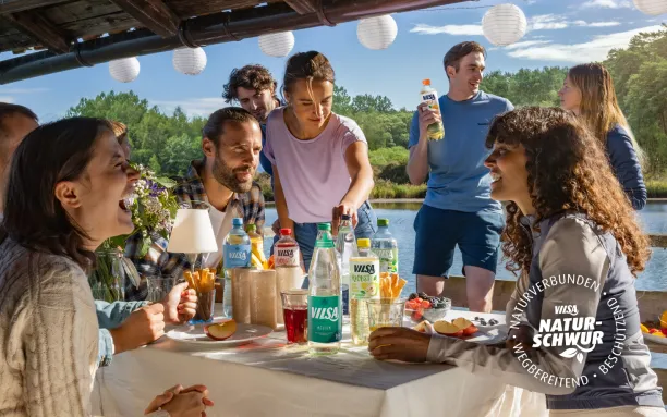 Eine Gruppe von Menschen sitzt an einem festlich gedeckten Tisch im Freien am Wasser. Sie lachen, genießen Getränke von VILSA und unterhalten sich. Im Hintergrund stehen weitere Personen, die ebenfalls in Gespräche vertieft sind.